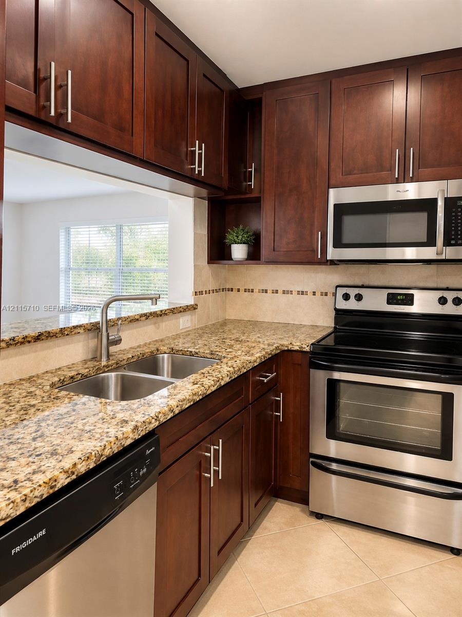 Kitchen featuring dark-toned cabinetry, stone-finish countertops, stainless steel appliances, ceramic tile backsplash, and a double-basin undermount sink