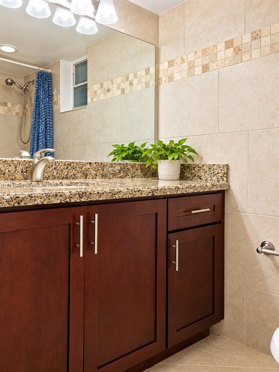 Vanity featuring dark wood-finish cabinetry, stone countertop, undermount sink, and polished metal faucet hardware, complemented by floor-to-ceiling wall tile with mosaic inlay and an expansive wall-mounted mirror