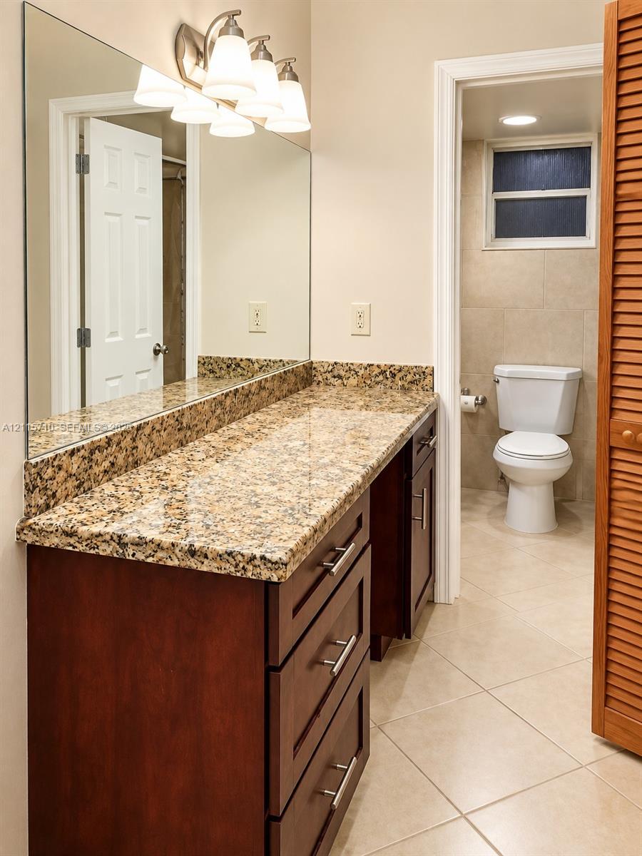 Vanity featuring stone-finish countertop, dark wood-finish cabinetry with brushed metal hardware, large wall-mounted mirror, multi-light fixture, and ceramic tile flooring
