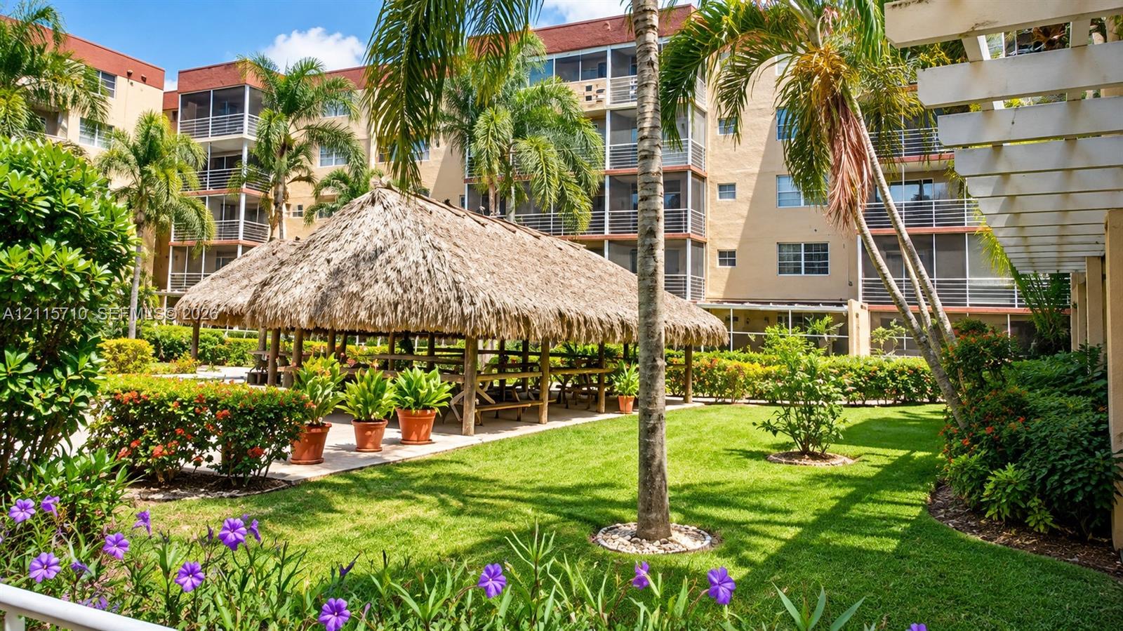 Manicured grounds featuring a thatched-roof pavilion, mature palm trees, decorative flowering shrubs, and multi-story building structures with screened-in balconies