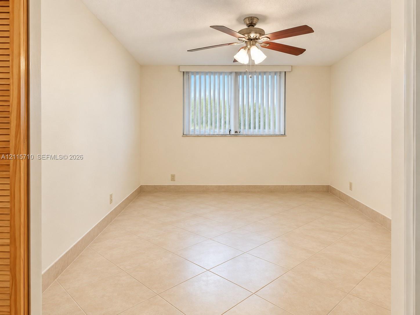 Interior room featuring large-format tile flooring, multi-blade ceiling fan with integrated light fixture, vertical window blinds, and built-in closet with louvered doors