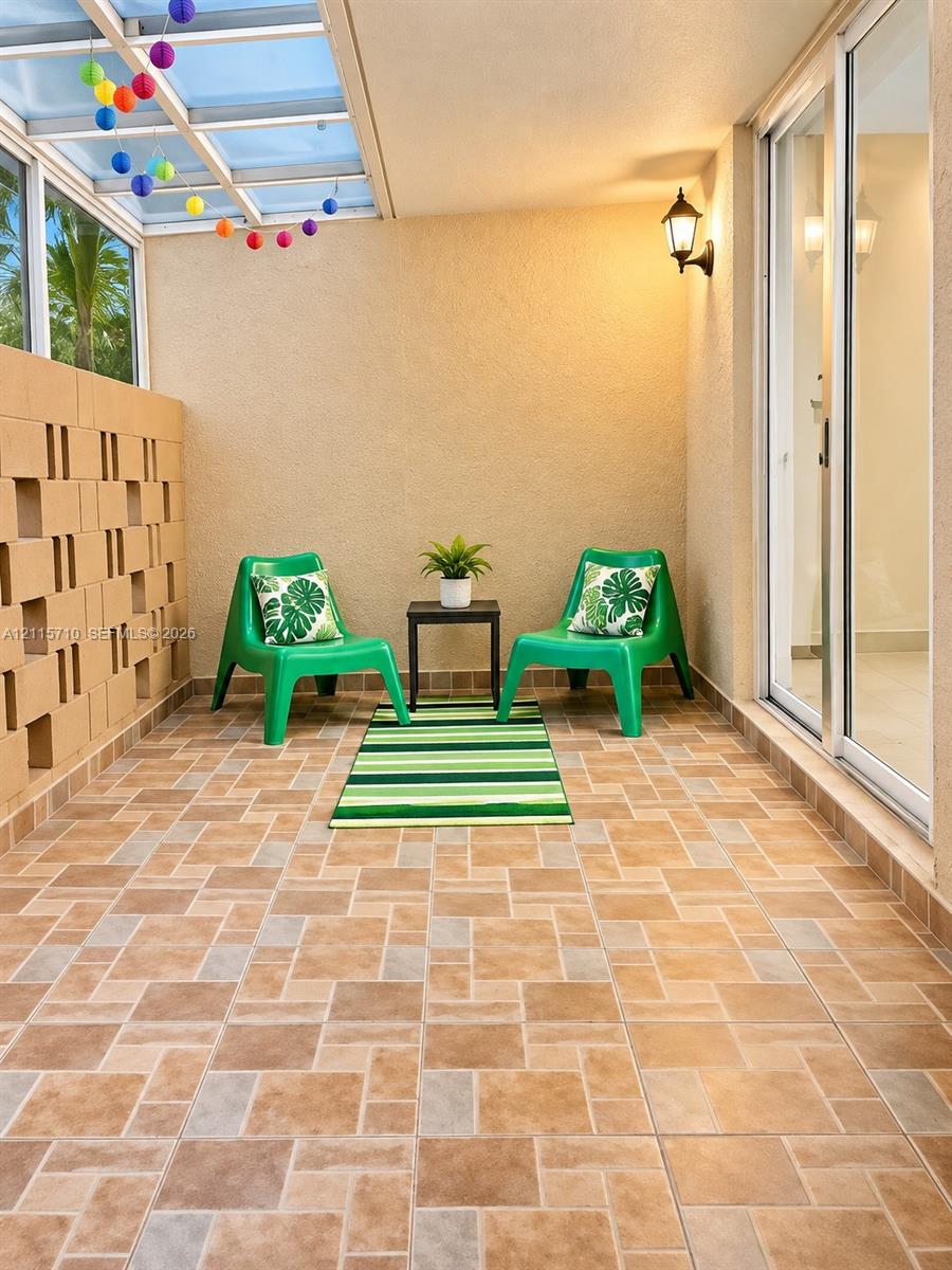 Covered patio featuring ceramic tile flooring, a geometric breeze block wall, a glass-paneled roof system, and wall-mounted exterior lighting