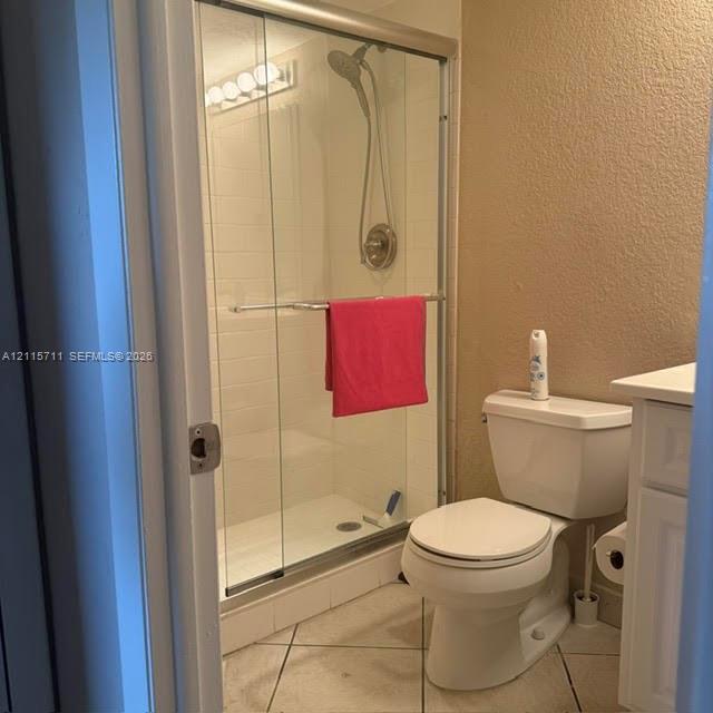 Walk-in shower with sliding glass door, adjustable handheld showerhead, white wall tile surround, white vanity, and ceramic tile flooring