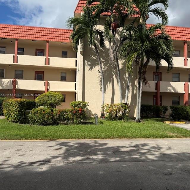 Multi-story building exterior featuring a textured stucco facade, a red tile roof, and multiple levels of covered balconies with vertical supports, set against a landscaped lawn with mature palm trees and manicured shrubbery