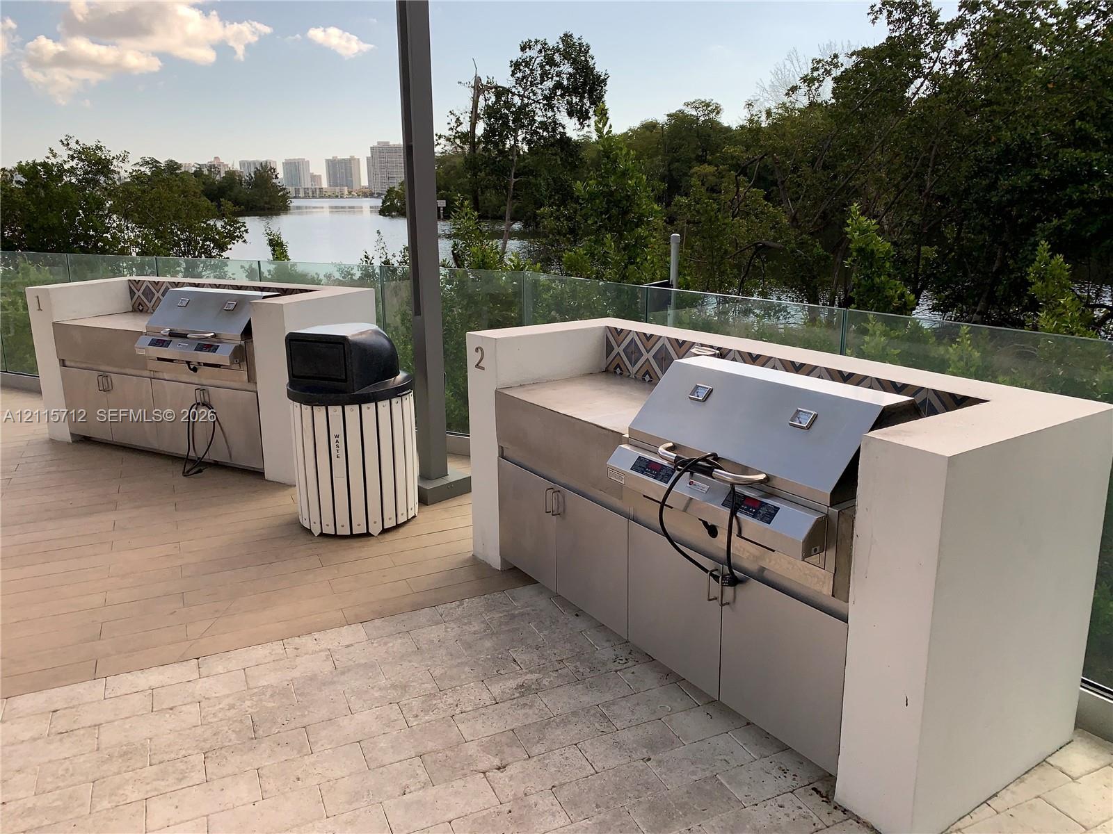 Outdoor kitchen featuring dual stainless steel built-in grills, concrete-surround masonry counters, decorative tile backsplashes, and glass panel railing