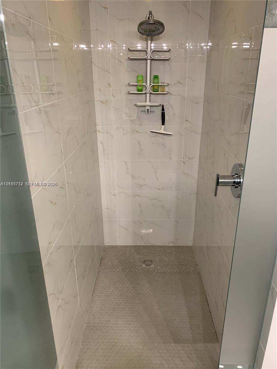 Walk-in shower featuring floor-to-ceiling large-format tile with marble-pattern veining, textured mosaic tile flooring, a wall-mounted rainfall showerhead, and a frameless glass enclosure