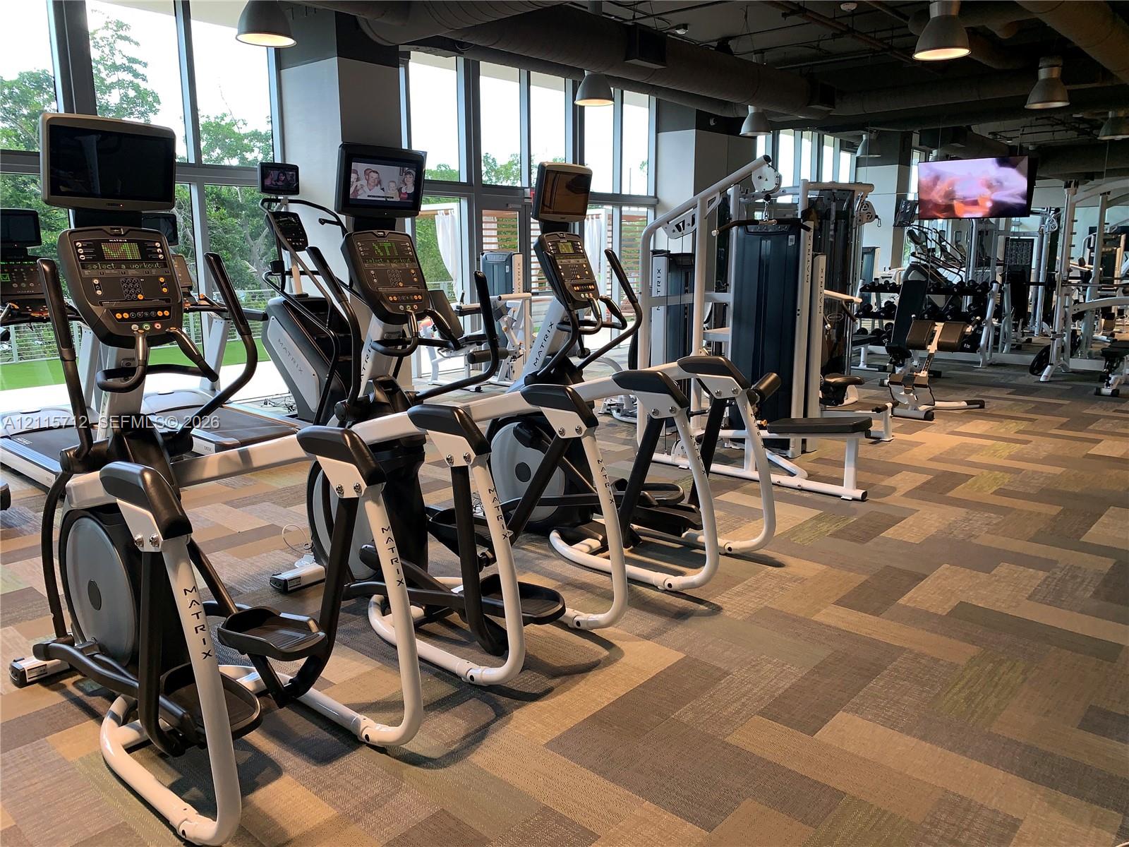 Fitness center featuring floor-to-ceiling windows, patterned carpet tile flooring, exposed industrial-style ceiling, and multi-station exercise equipment
