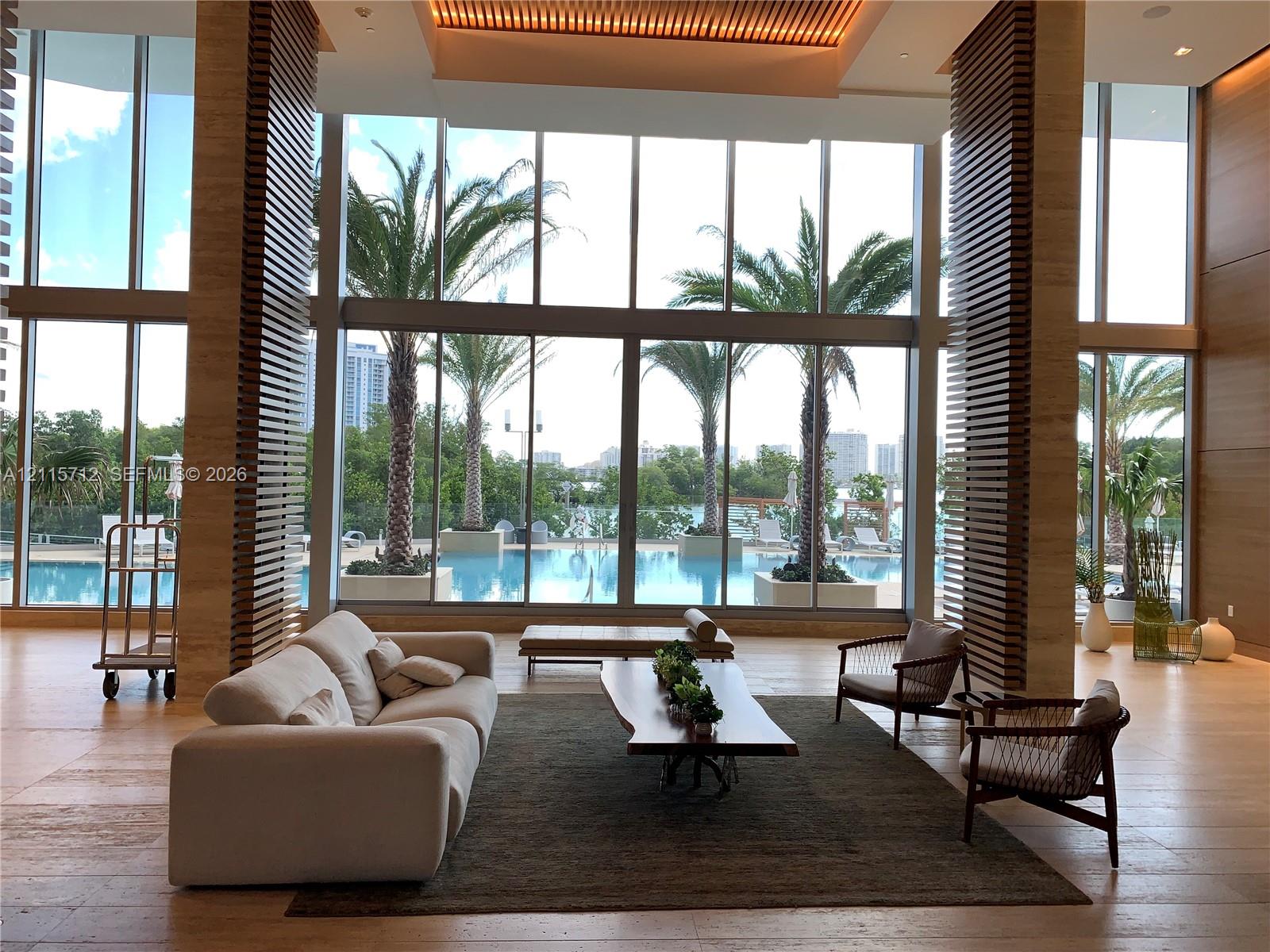 Double-height floor-to-ceiling windows, stone flooring, decorative slatted wood-finish columns, recessed architectural ceiling lighting, and exterior swimming pool