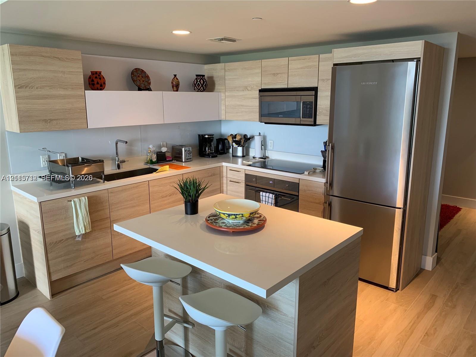 Modern kitchen featuring wood-finish cabinetry, white solid-surface countertops, a center island with bar seating, stainless steel appliances, and integrated recessed lighting