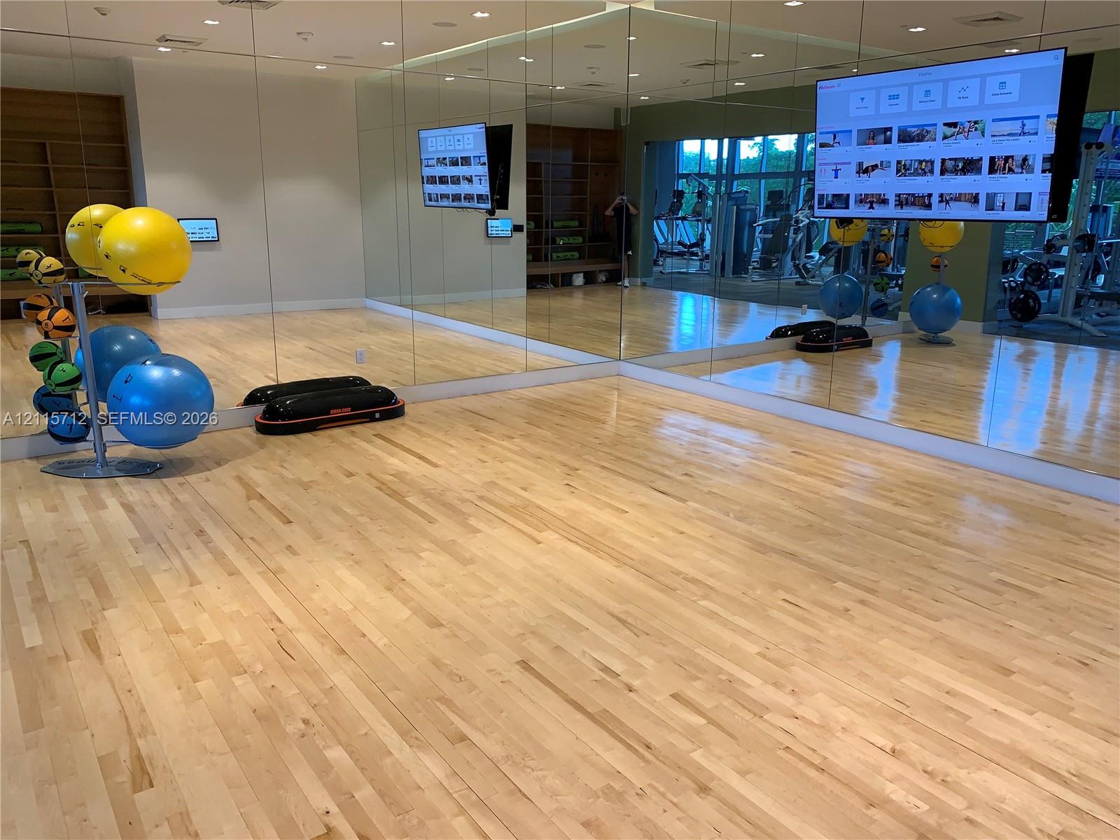 Fitness studio featuring wood-finish flooring, floor-to-ceiling mirrored walls, recessed lighting, and wall-mounted display monitors