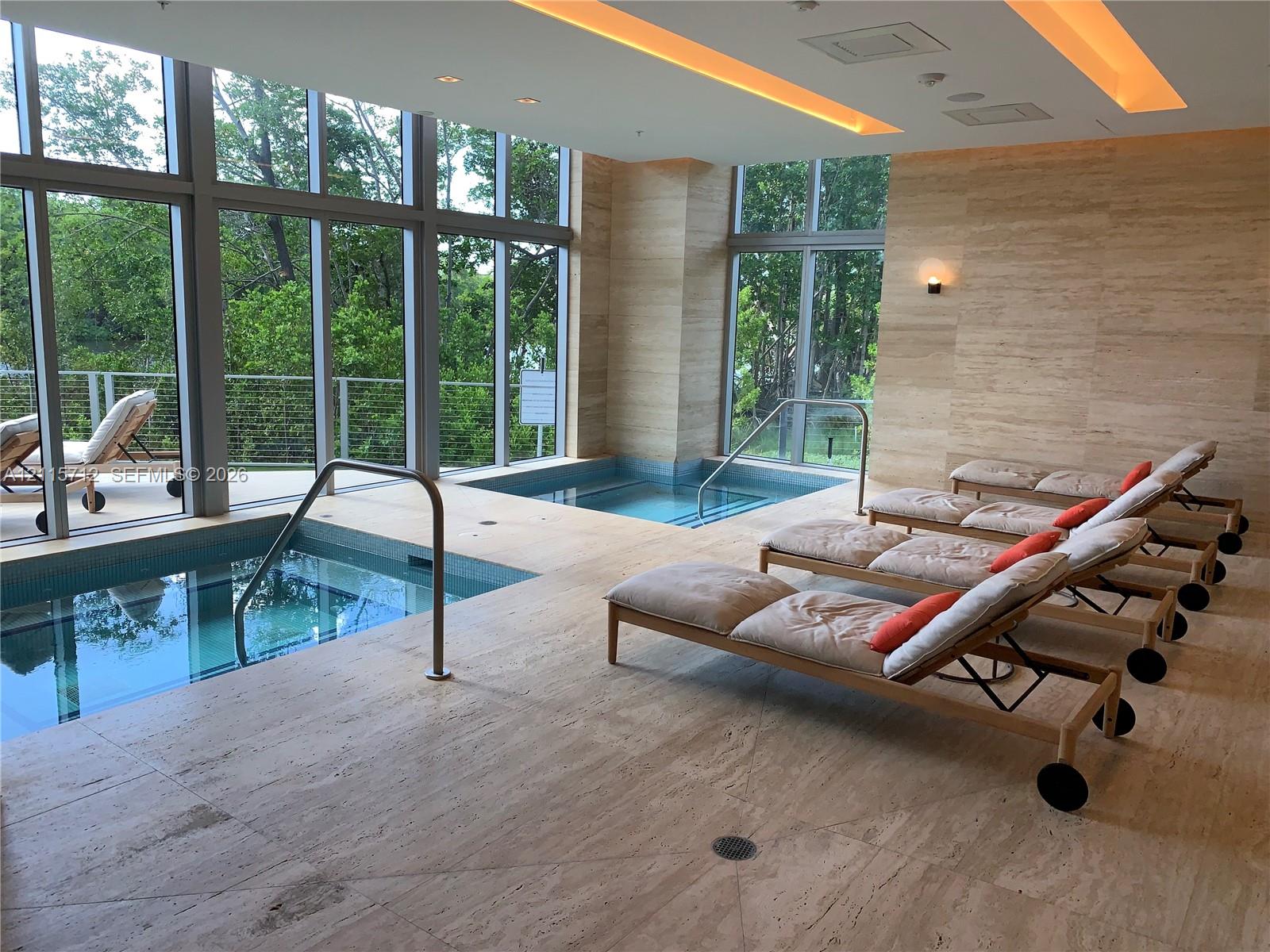 Indoor spa area featuring dual tiled soaking pools, full-height floor-to-ceiling windows, recessed cove lighting, and stone-tiled walls and flooring