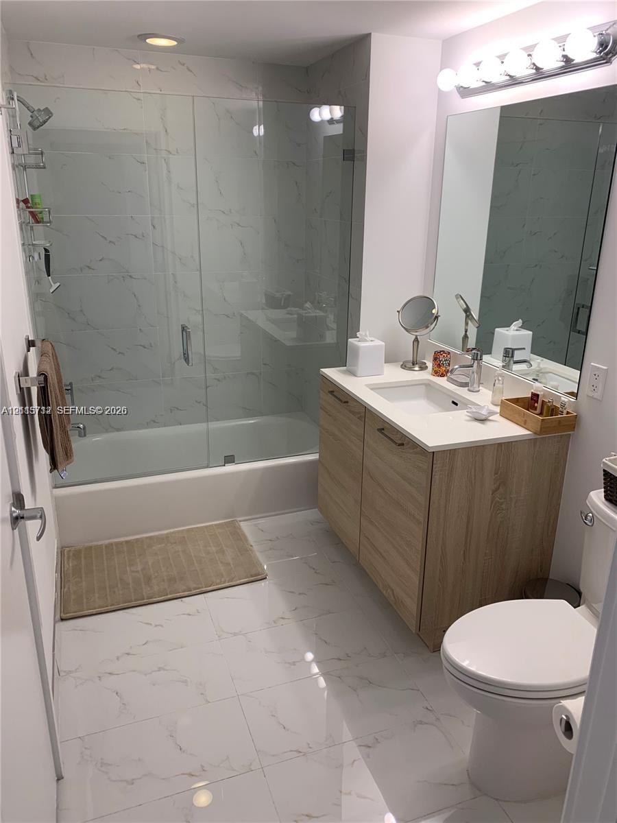Bathroom featuring a wood-finish vanity, solid-surface countertop with integrated sink, glass-enclosed shower and tub combination with tile surround, and polished tile flooring