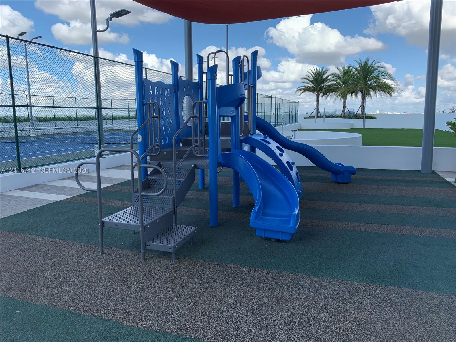 Playground structure featuring multiple slides and integrated staircases set on a rubberized surface, covered by a permanent overhead canopy, with an enclosed court area