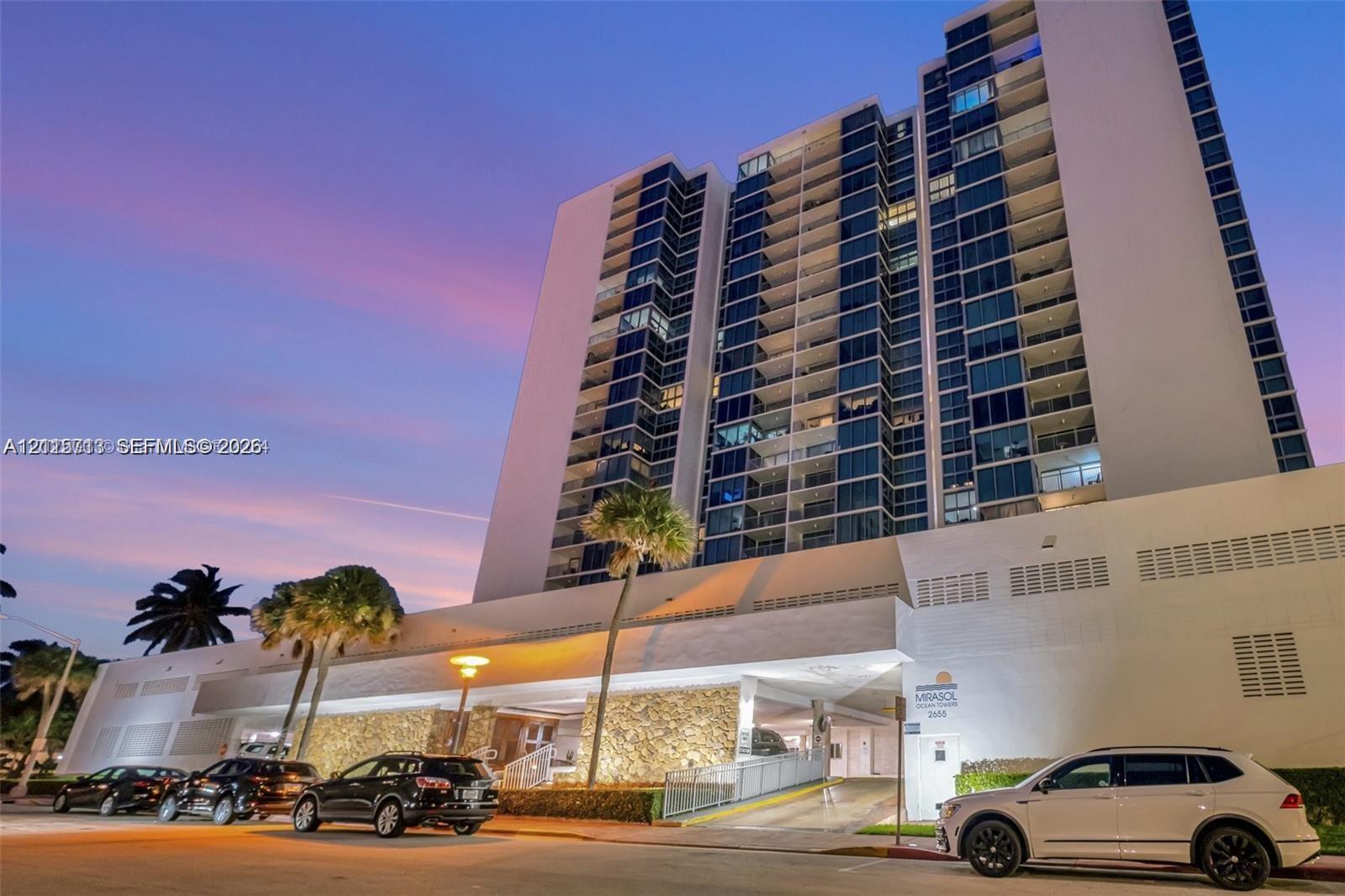 High-rise residential exterior featuring expansive glass paneling, recessed private balconies, stone veneer podium cladding, and integrated ground-level parking