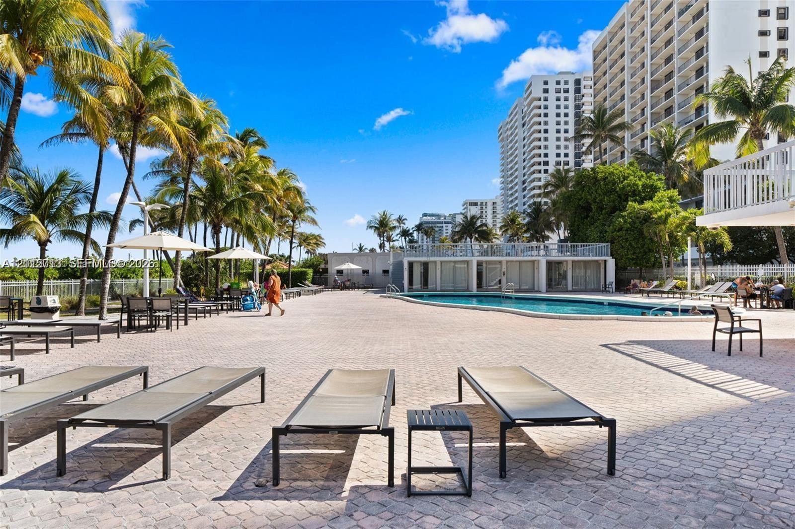 Outdoor pool area featuring a paved sun deck, mature palm trees, and a structure with floor-to-ceiling windows