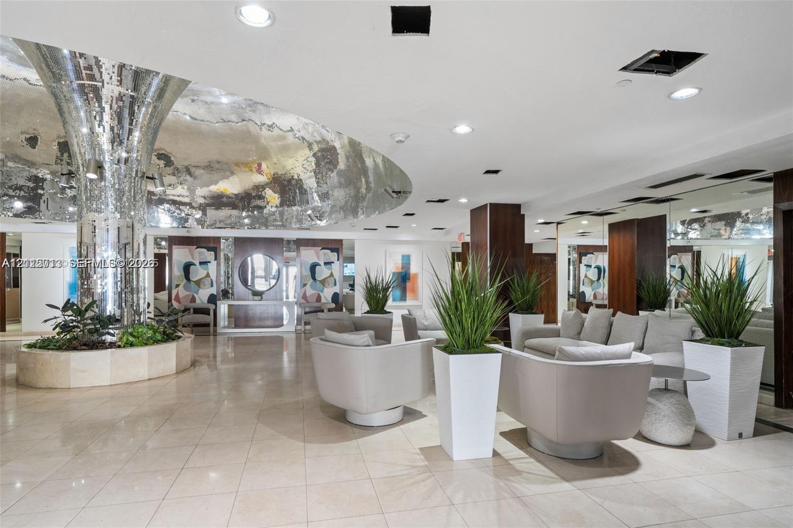 Grand lobby featuring polished stone flooring, a cylindrical mirrored pillar, a circular recessed ceiling element with reflective finish, and wall-to-wall mirrored paneling