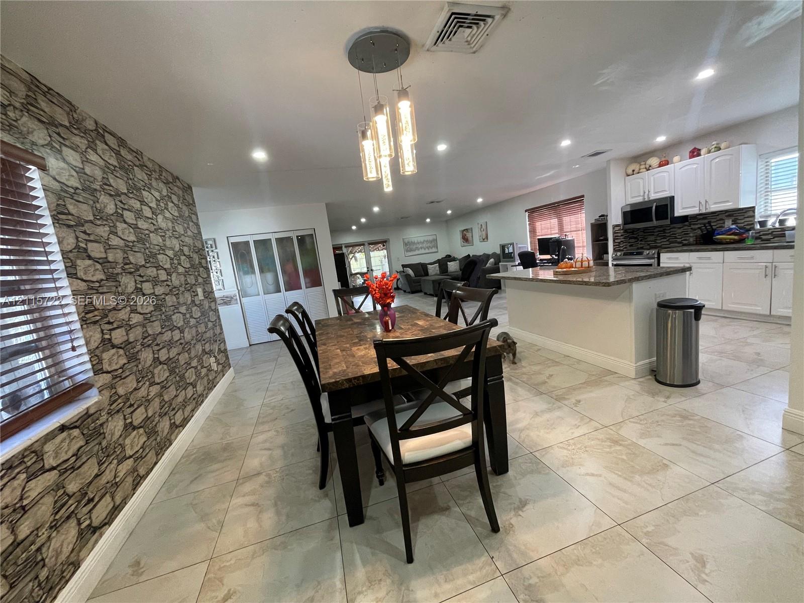 Stone-textured accent wall, tile flooring, kitchen island with a stone-finish countertop, white shaker-style cabinetry, and a multi-pendant light fixture