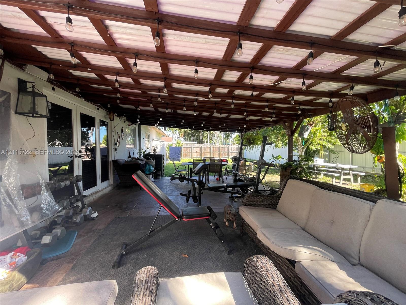 Covered outdoor patio featuring a wood-beam pergola structure, tiled flooring, mounted string lighting, and an attached wall-mounted fan