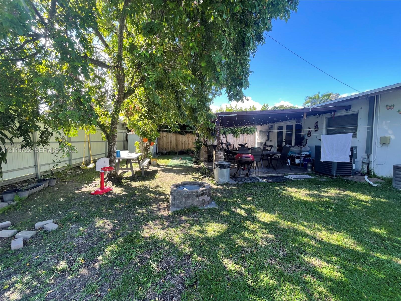 Backyard featuring a stone-encased fire pit, covered patio, mature canopy trees, and perimeter fencing