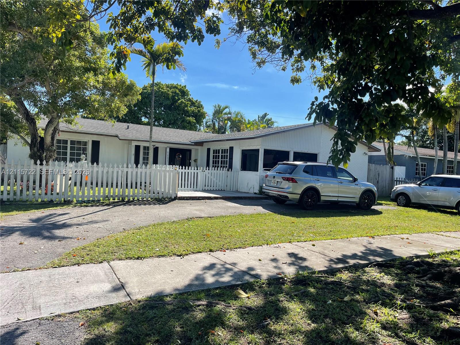 Single-story residence featuring a white exterior, picket fencing, mature shade trees, and a screened-in porch area