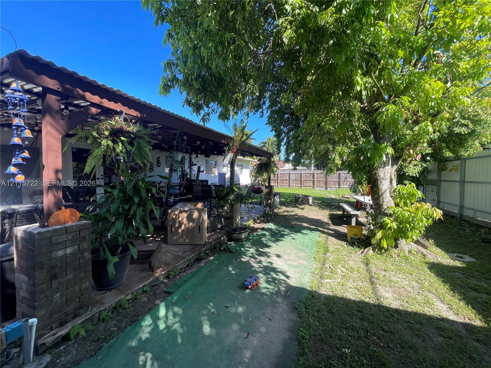 Covered patio featuring a timber-frame structure and brick masonry pillar, situated within a backyard defined by mature trees and perimeter wood fencing