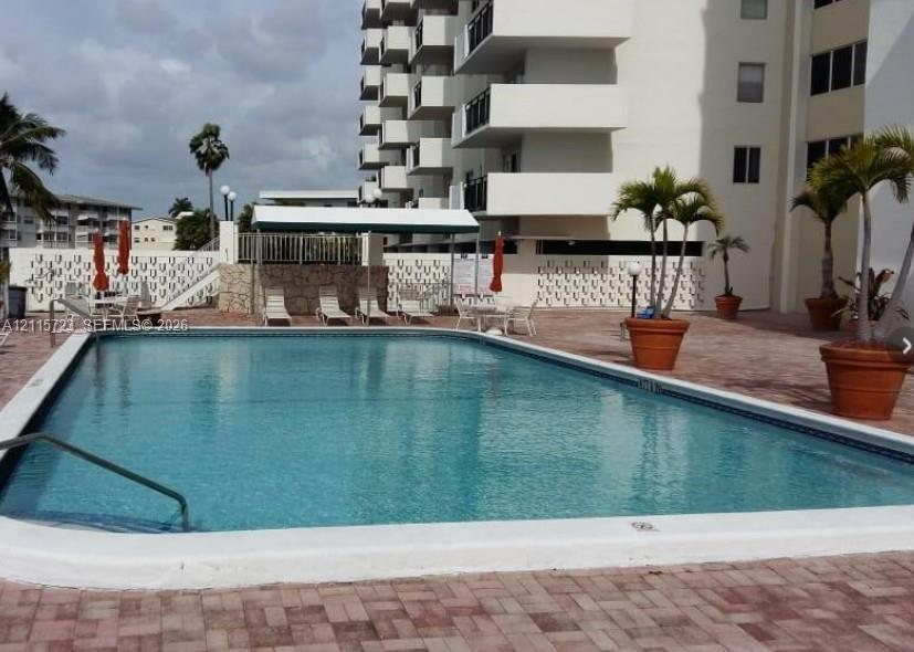 Rectangular swimming pool featuring a brick-paver deck, stone-accented retaining wall, covered poolside canopy, and multi-story high-rise exterior