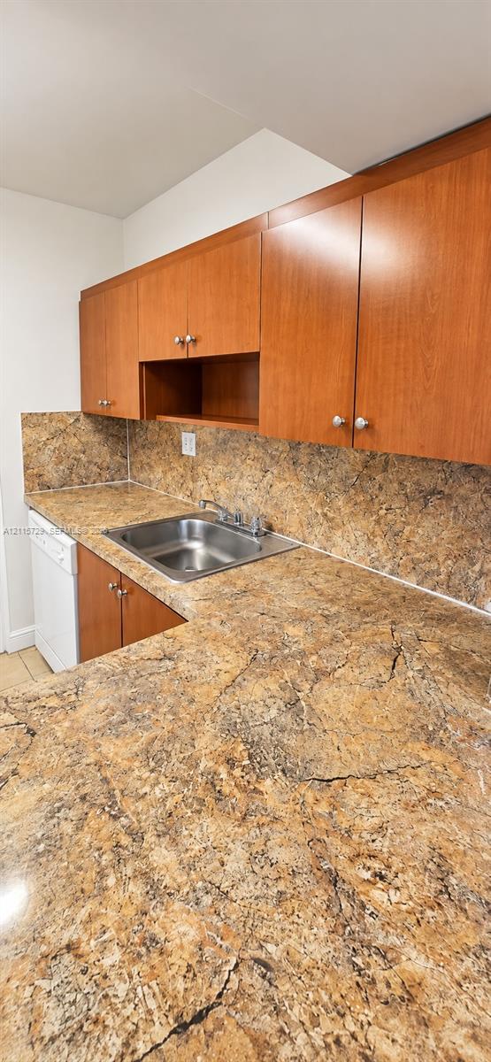 Kitchen featuring wood-finish cabinetry, stone-patterned countertops, matching stone-patterned backsplash, stainless steel drop-in sink with a chrome faucet, and a white built-in dishwasher Kitchen featuring wood-finish cabinetry, stone-patterned countertops, matching stone-patterned backsplash, stainless steel drop-in sink with a chrome faucet, and a white built-in dishwasher