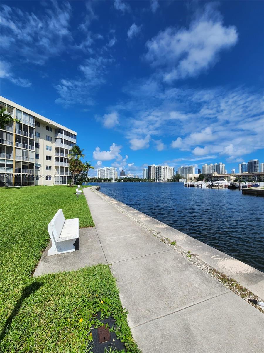 Waterfront grounds featuring a concrete bulkhead, paved walkway, manicured lawn, and fixed stone seating