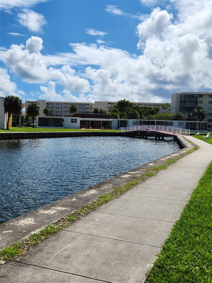 Waterfront grounds featuring a concrete walking path, a pedestrian footbridge, manicured lawn, and mature palm trees