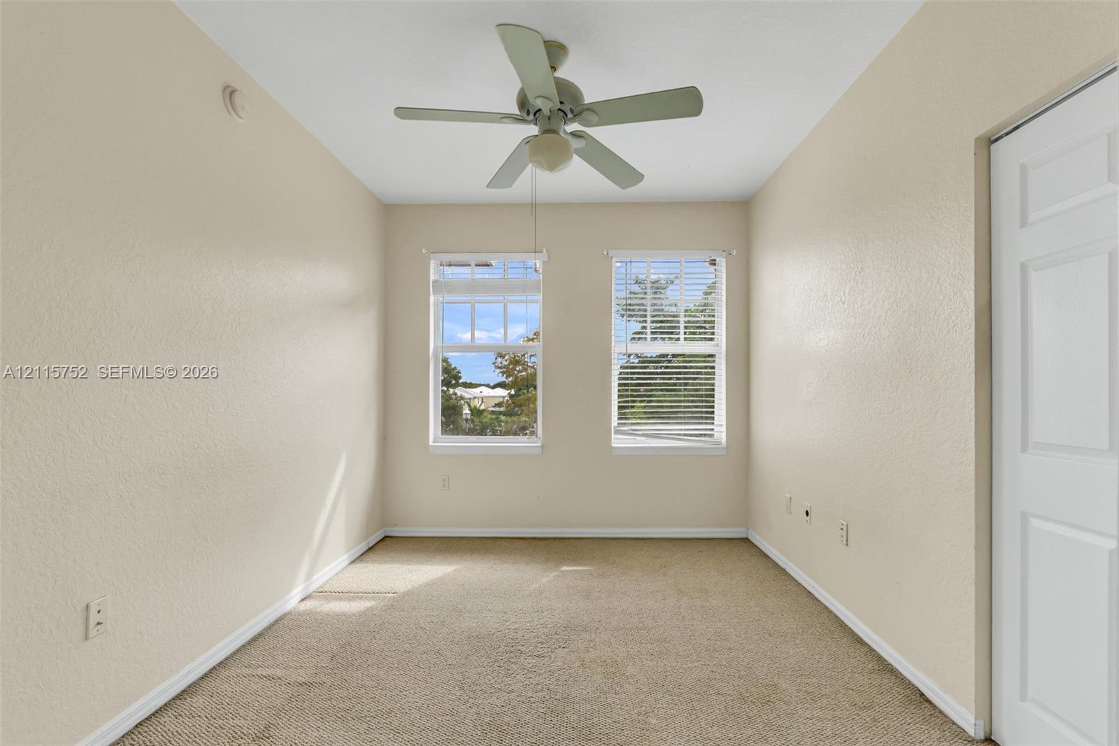 Neutral-toned interior space featuring carpeting, dual windows with blinds, a ceiling fan with integrated lighting, and a paneled closet door