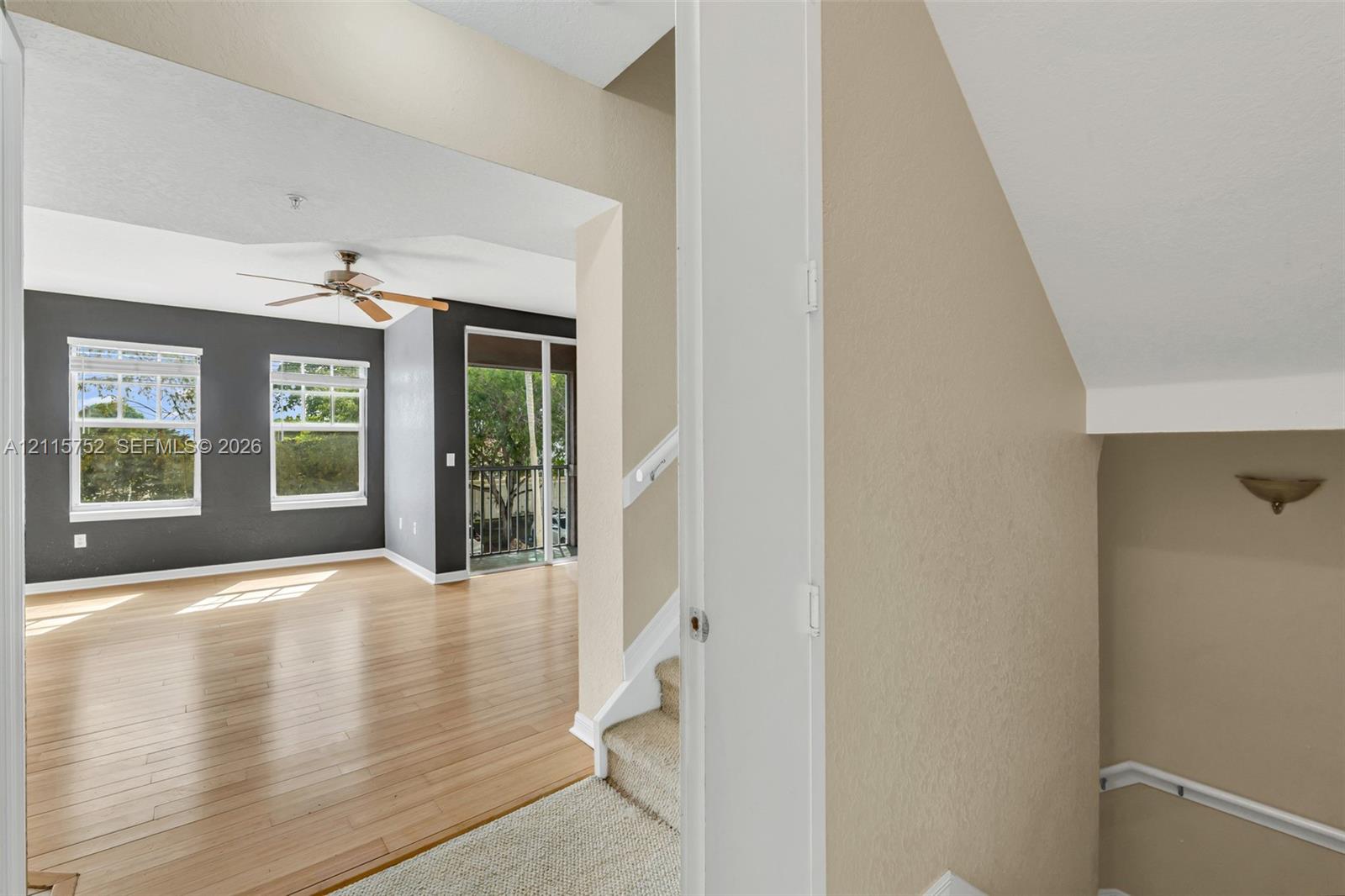 Living area featuring wood-finish flooring, charcoal-toned accent wall, ceiling fan, and large windows with exterior access