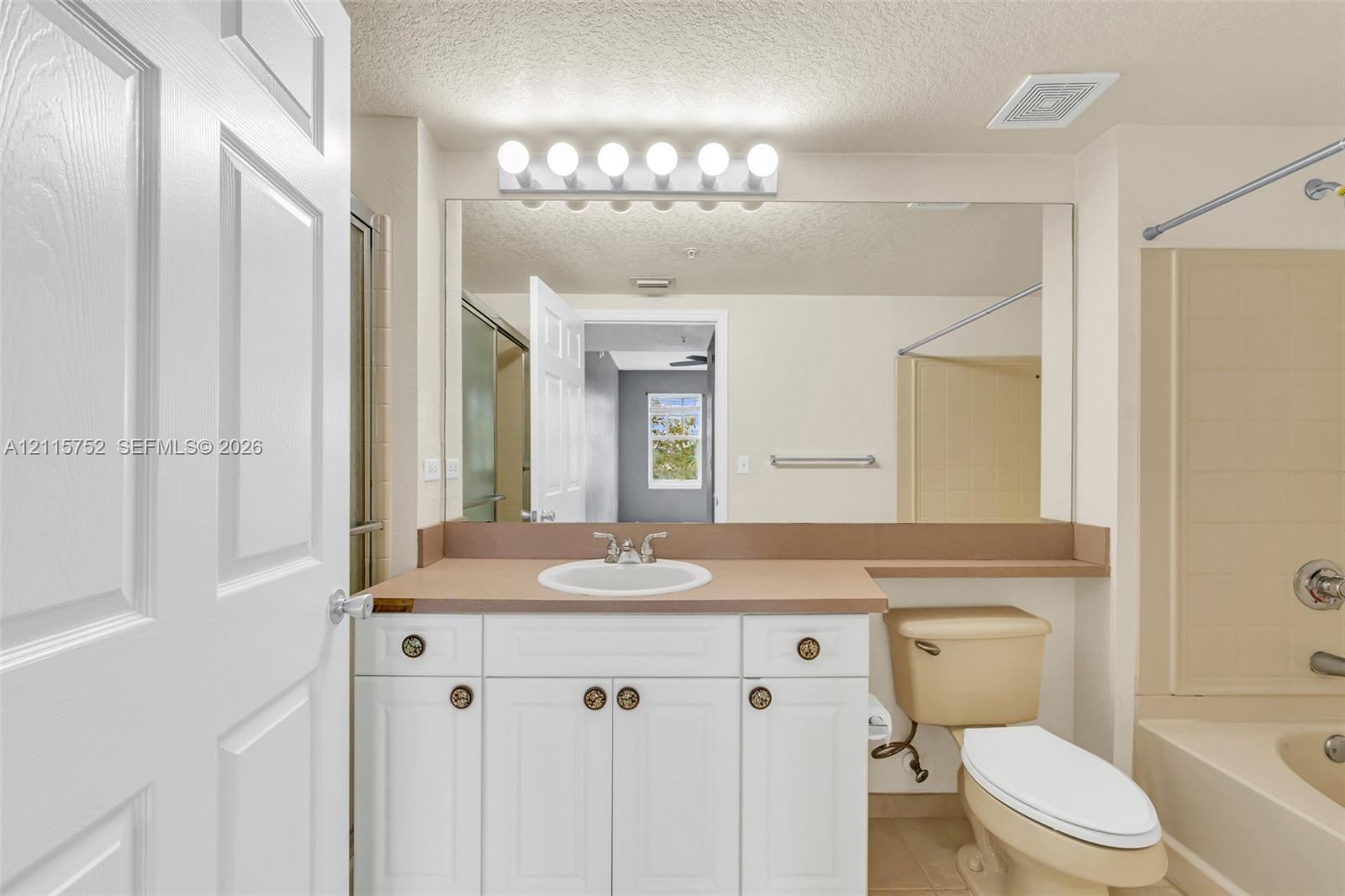 Bathroom featuring white cabinetry, solid-surface countertop with integrated basin, full-width mirror, six-light vanity fixture, and a tub-shower combination