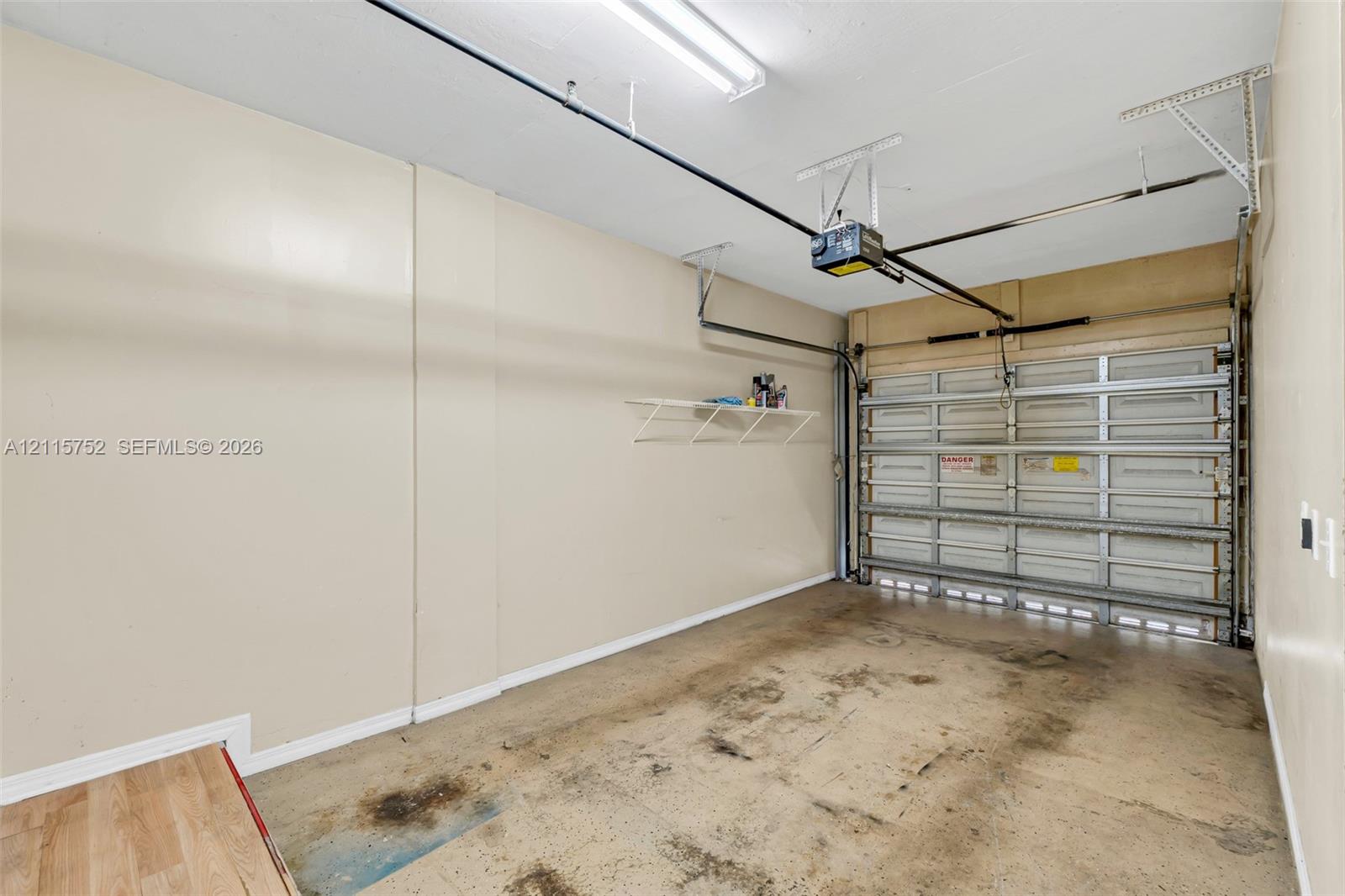 Enclosed garage featuring a sectional metal door, automated door opener, wall-mounted wire shelving, and overhead fluorescent lighting Enclosed garage featuring a sectional metal door, automated door opener, wall-mounted wire shelving, and overhead fluorescent lighting