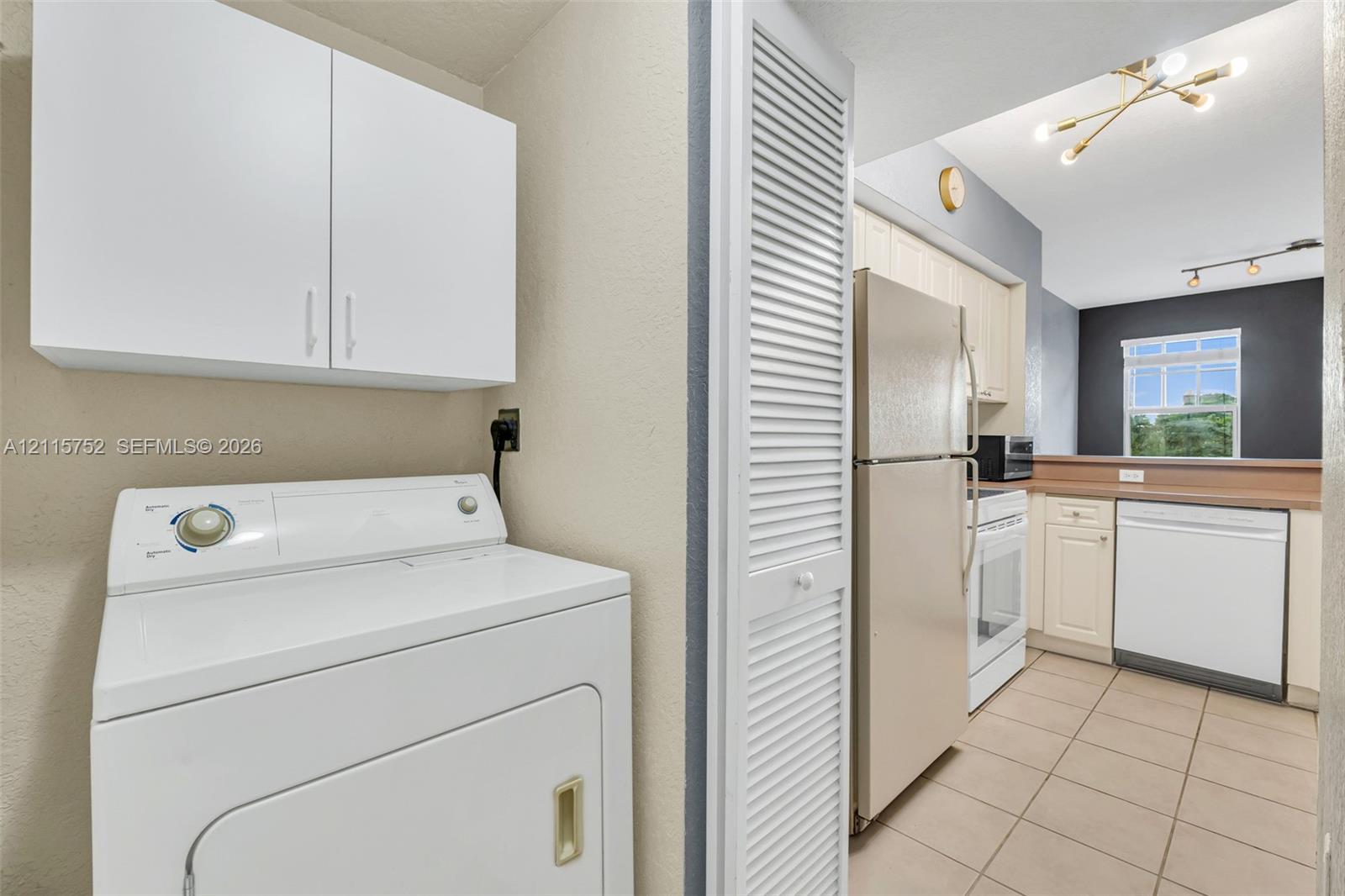 Laundry area featuring white wall-mounted cabinetry and louvered closet doors, leading to a kitchen equipped with tile flooring, light-toned cabinetry, and a modern multi-bulb overhead light fixture