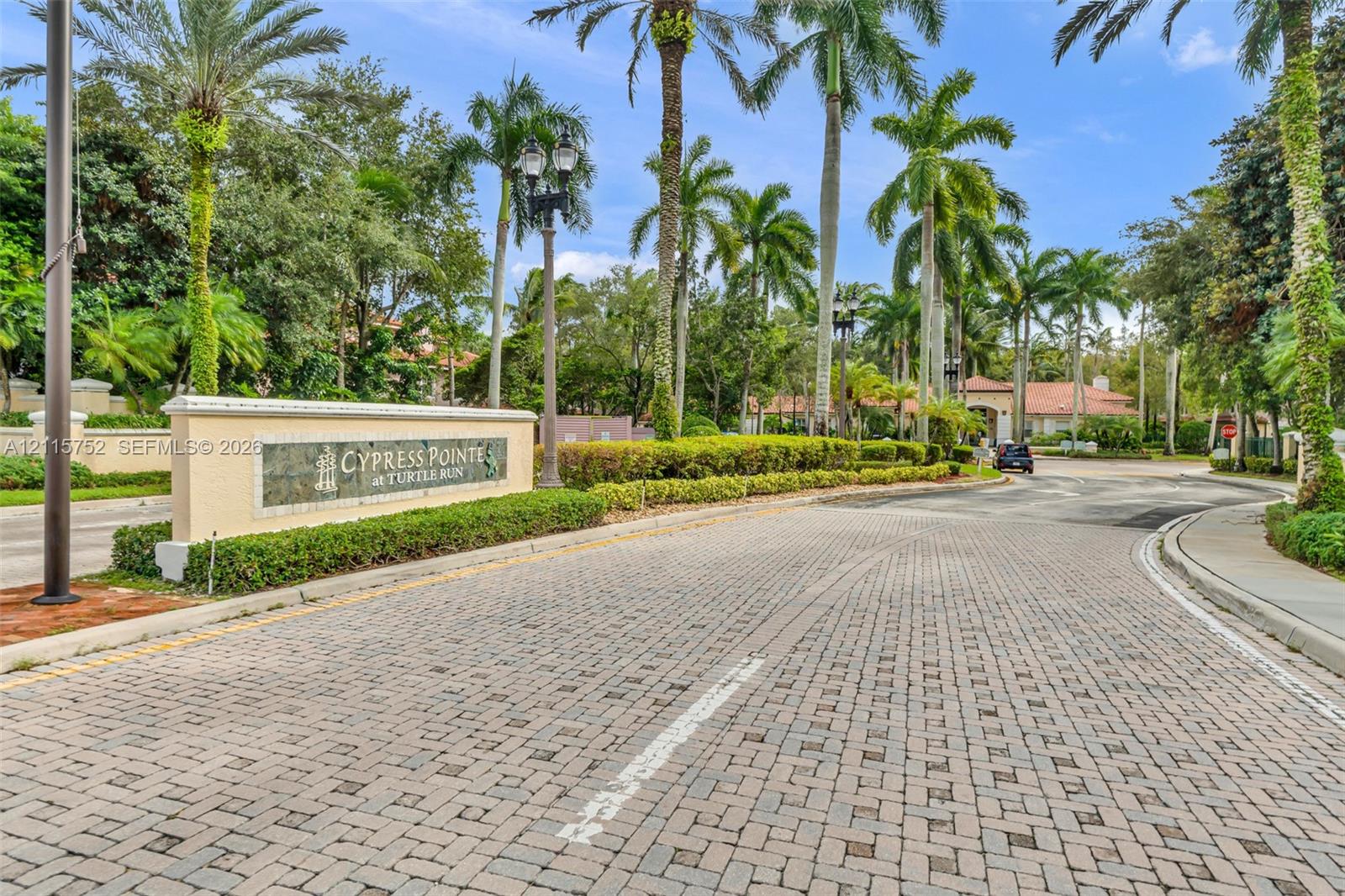 Community entrance featuring decorative stone signage, paver road surface, mature palm trees, formal landscaping, and iron-style lighting fixtures