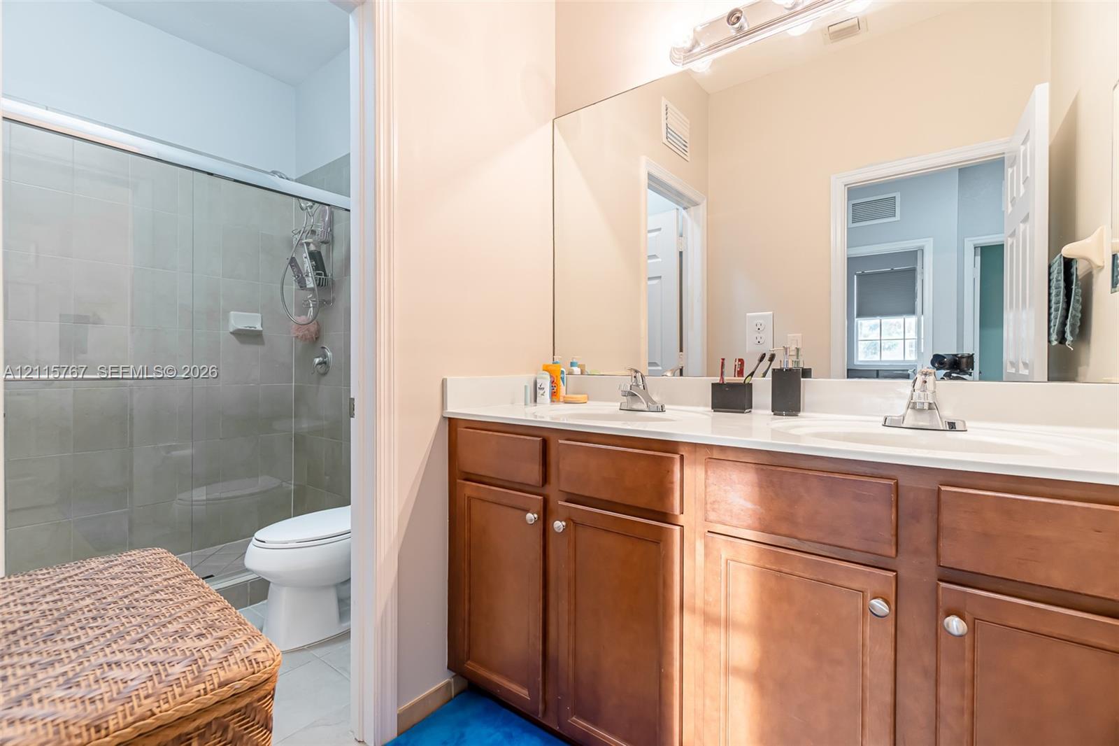 Dual-sink vanity with wood-finish cabinetry, solid surface countertops, glass-enclosed shower, and full-width vanity mirror
