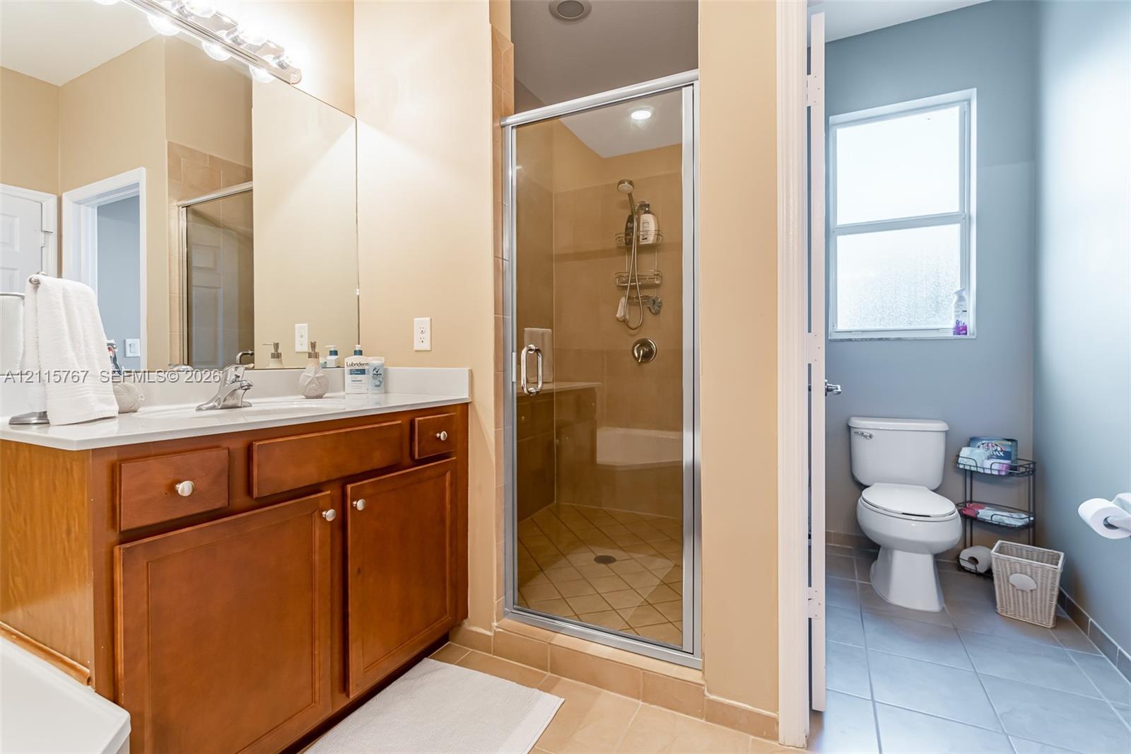 Bathroom featuring a wood-finish vanity with a light-toned countertop, a shower with tile surround and glass enclosure, tile flooring, and a window