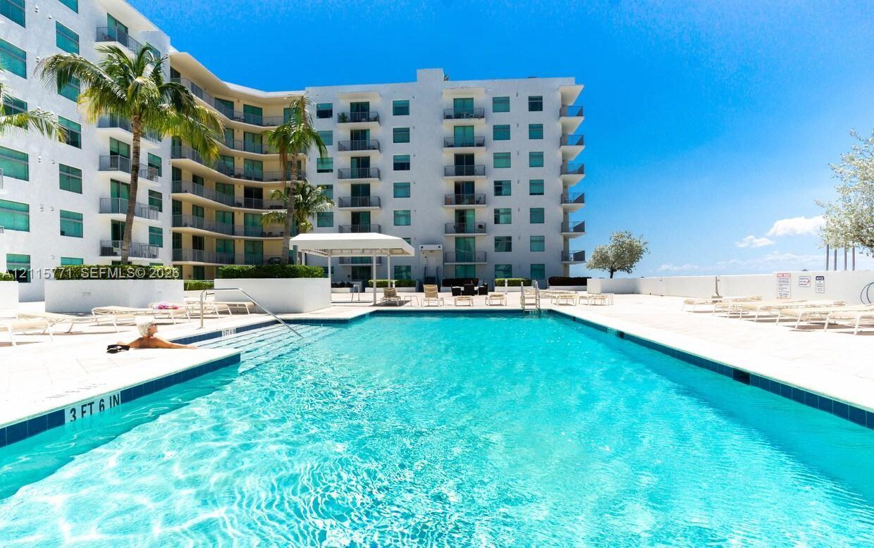 Swimming pool with built-in stairs, surrounded by a paved deck, palm trees, and multi-story building exterior