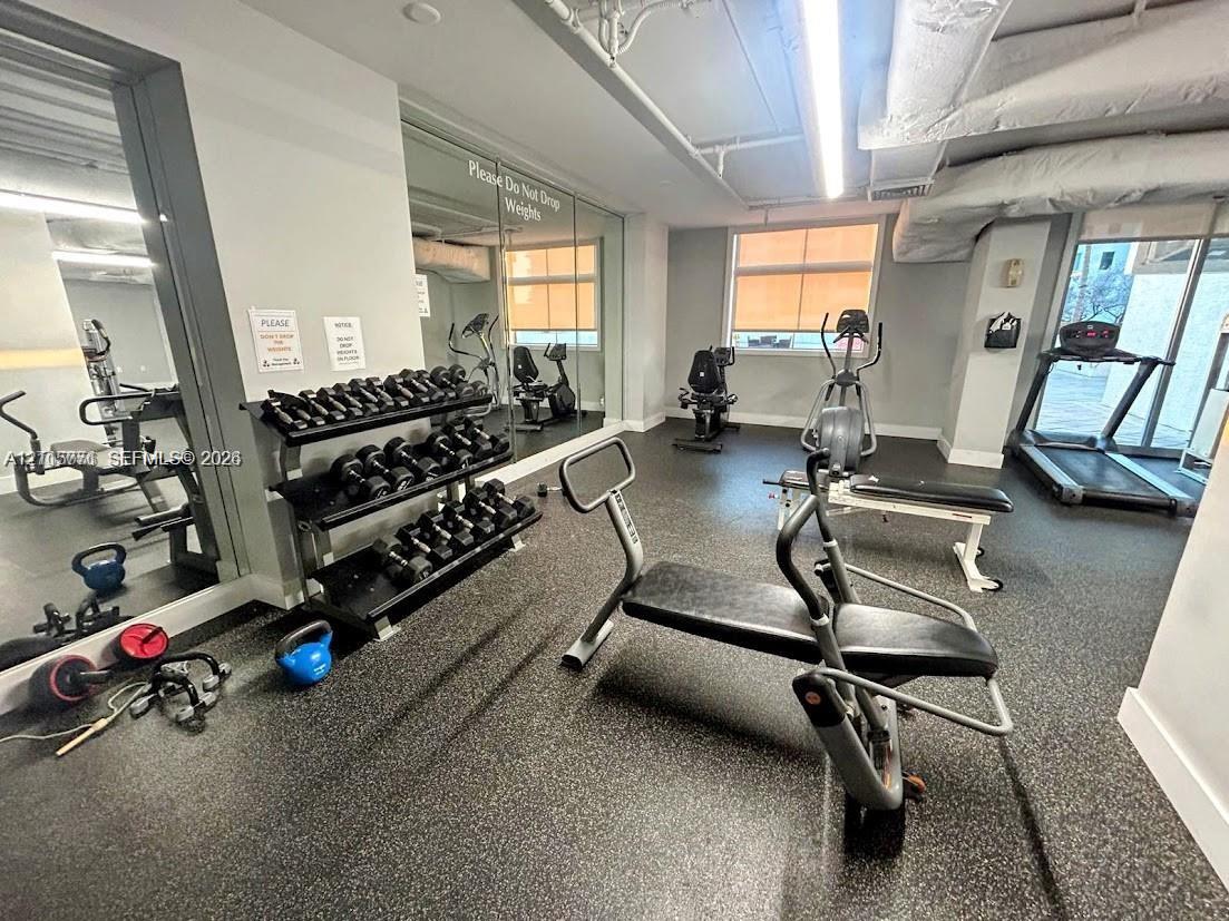 Fitness facility featuring rubberized flooring, expansive wall-to-wall mirrors, industrial-style exposed ceiling, and large windows with roller shades