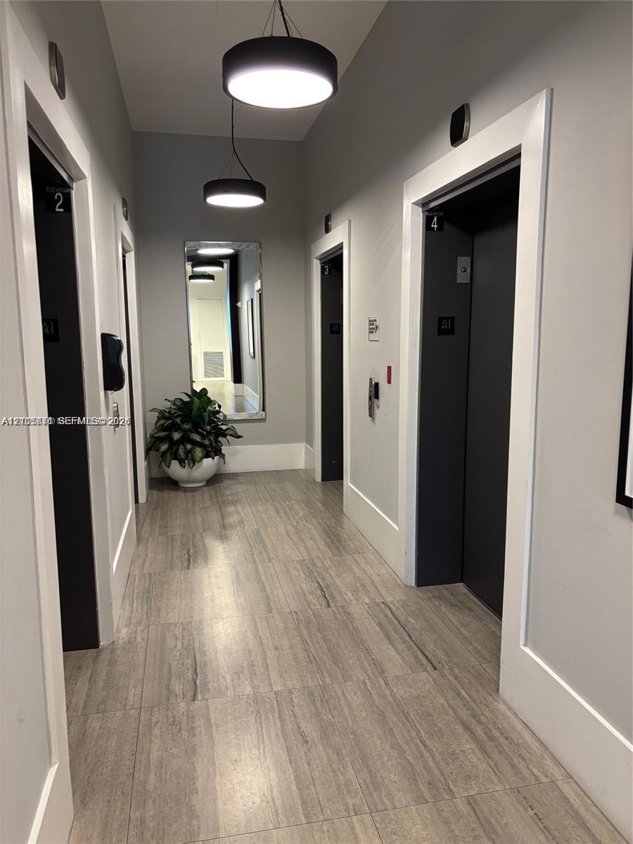 Hallway featuring wood-finish tile flooring, matte black elevator doors, wall-mounted modern pendant lighting, and a recessed wall mirror
