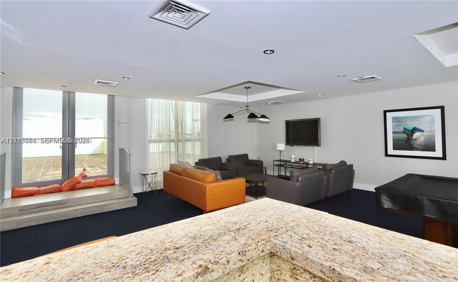 Expansive interior featuring high ceilings with recessed lighting, stone-surface countertop, ceiling-mounted multi-arm light fixture, floor-to-ceiling windows, and a raised platform with an integrated glass railing
