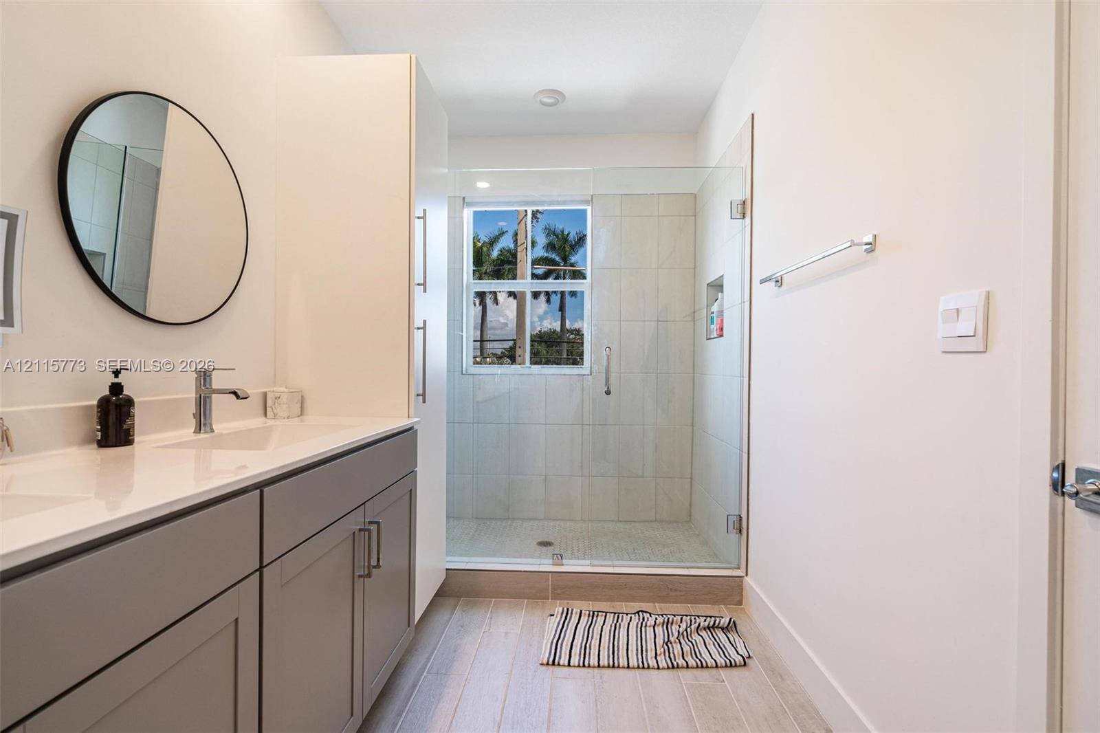 Bathroom featuring a neutral-toned vanity with a solid-surface countertop, glass-enclosed walk-in shower with tiled surround, wood-finish flooring, and a circular wall-mounted mirror