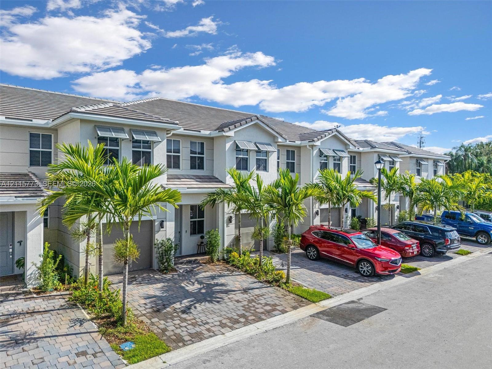 Multi-unit residential exterior featuring a light-toned stucco faade, shingle roofing, integrated garage doors, and paver driveways lined with palm trees