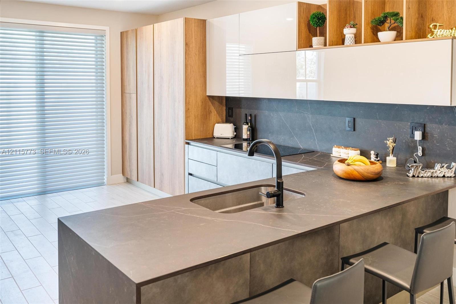 Kitchen featuring two-tone cabinetry with wood-finish vertical panels and white upper cabinets, expansive dark stone-finish countertops, undermount stainless steel sink, matte black gooseneck faucet, and light-toned tile flooring