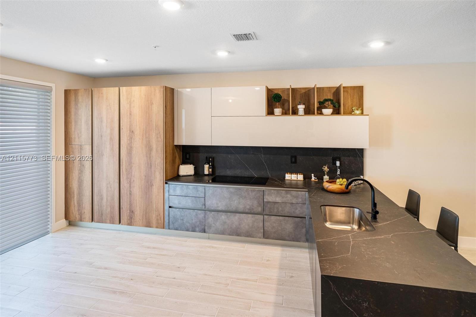 Kitchen featuring flat-panel wood-finish cabinetry, gloss-finish upper cabinets, stone-surfaced countertops, integrated sink with matte black faucet, and stone-surfaced backsplash