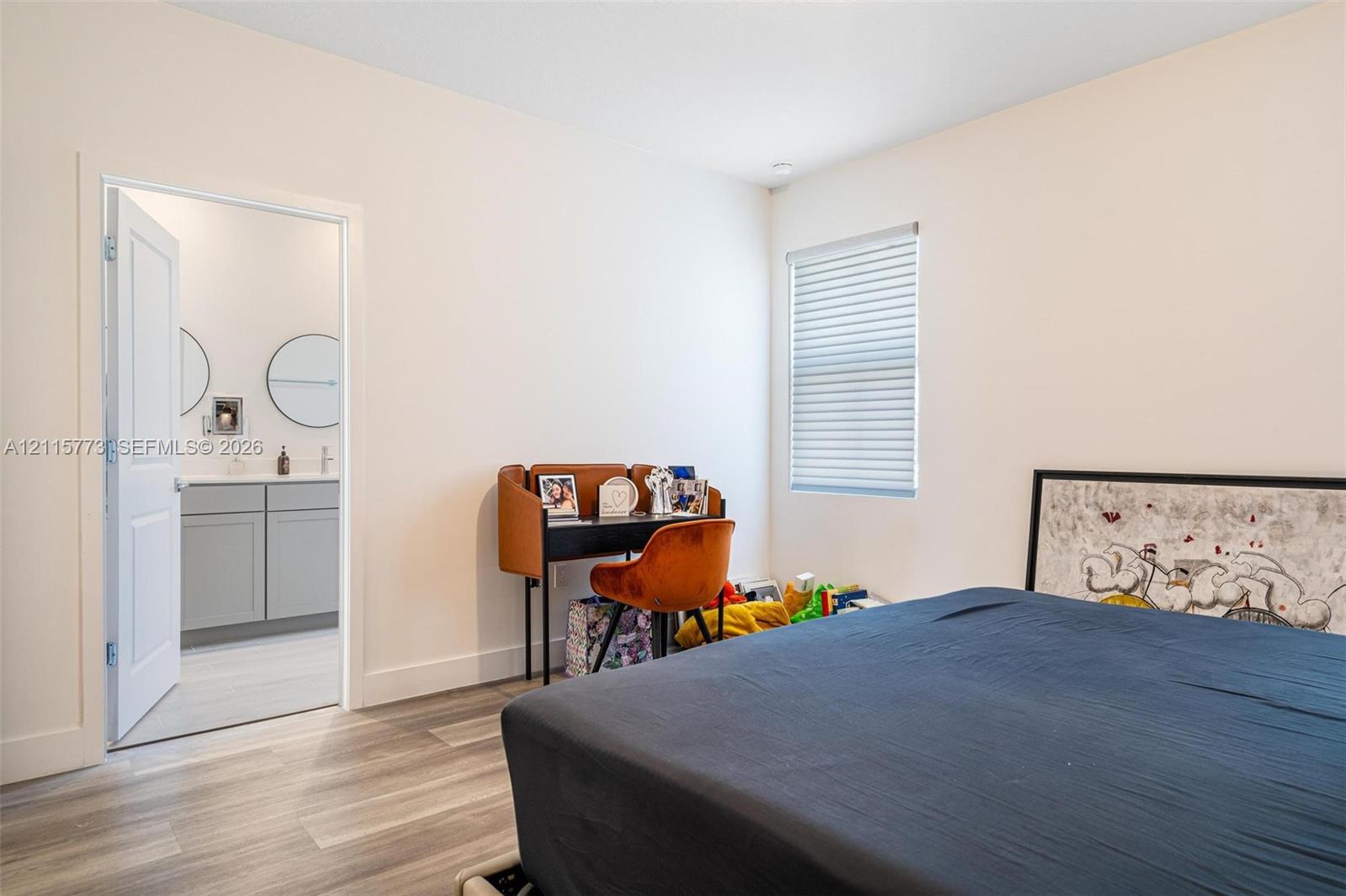 Neutral-toned room featuring wood-finish flooring, horizontal window blinds, and an interior door opening to a bathroom with a gray dual-sink vanity