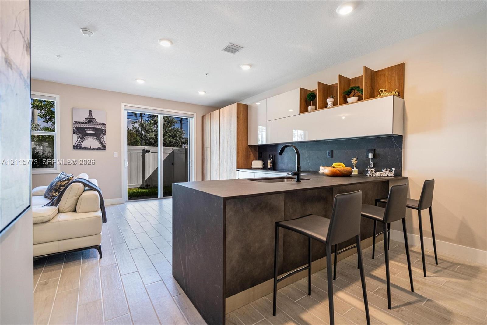 Kitchen featuring a waterfall-edge island with matte dark countertops, two-tone cabinetry, recessed lighting, and wood-finish flooring throughout Kitchen featuring a waterfall-edge island with matte dark countertops, two-tone cabinetry, recessed lighting, and wood-finish flooring throughout