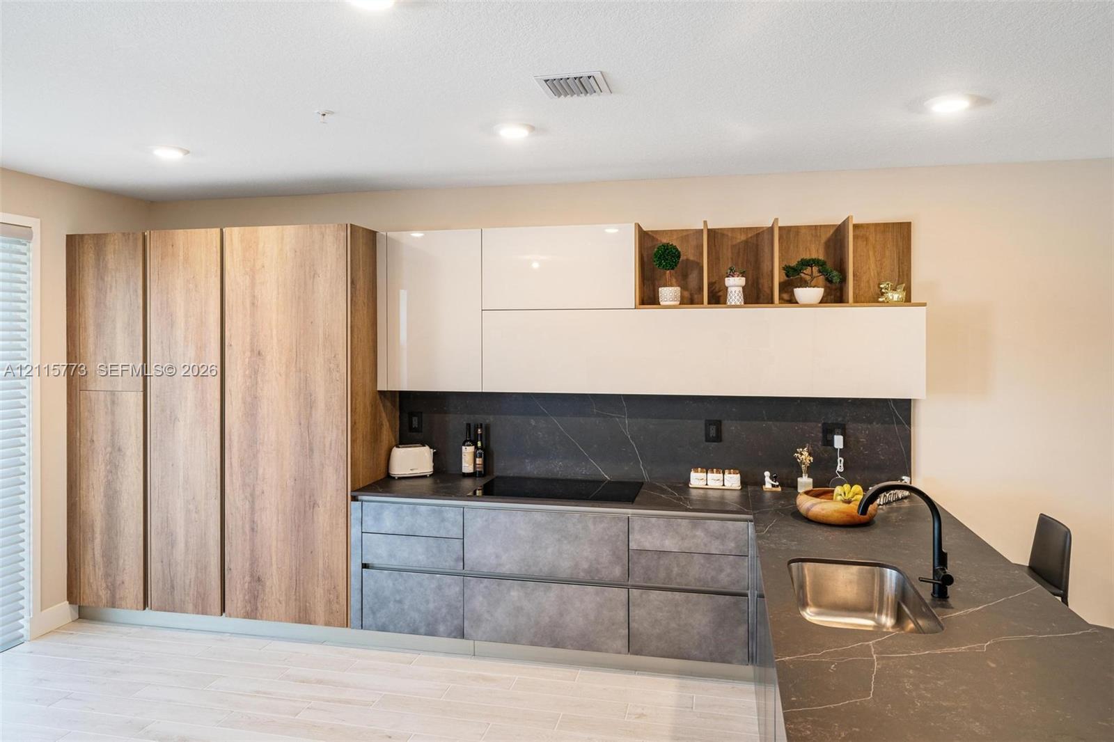 Kitchen featuring high-gloss white upper cabinetry, integrated wood-finish full-height storage, matte stone-texture lower cabinetry, stone-finish countertops, and an undermount stainless steel sink with a matte black faucet