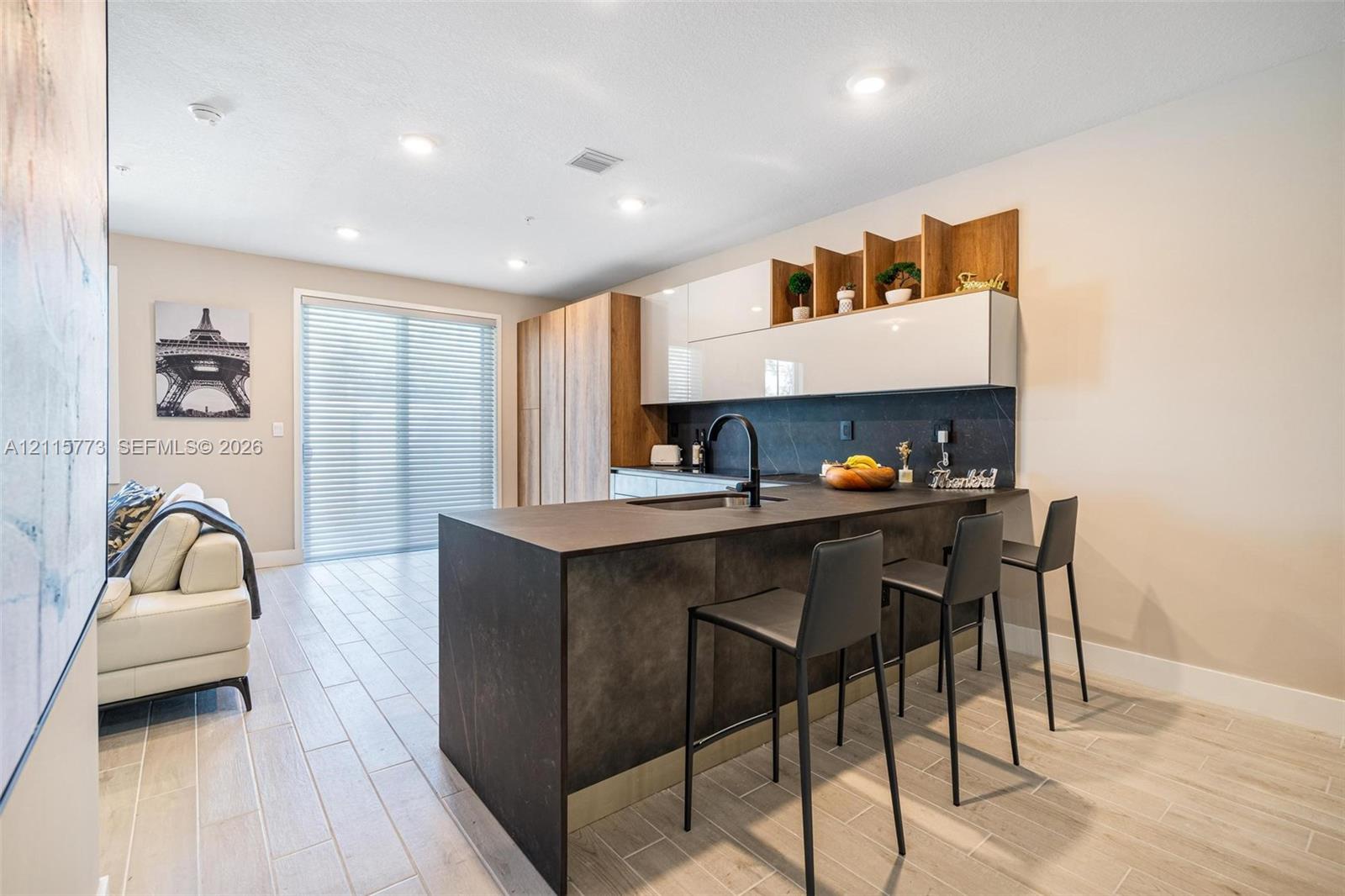 Kitchen featuring a matte dark finish island with a built-in sink, high-gloss white cabinetry, integrated wood-tone shelving, stone-finish backsplash, and light-toned wood-finish flooring