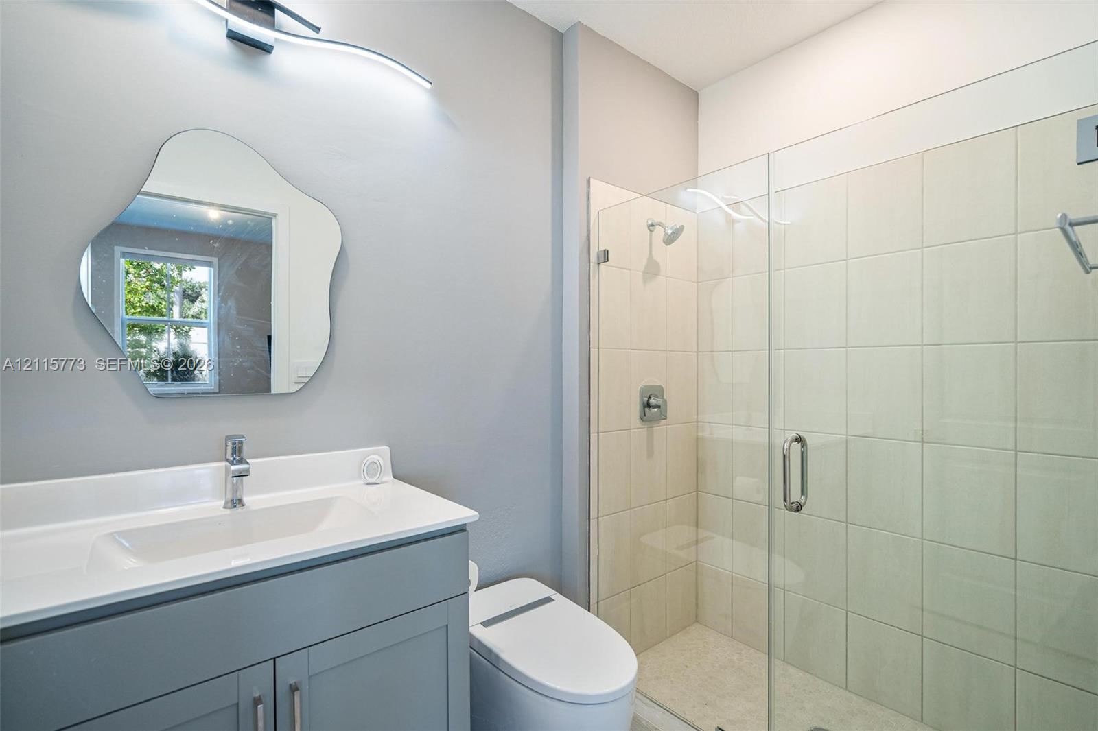 Grey vanity with a white countertop and integrated sink, chrome faucet, wall-mounted curved light fixture, organic-shaped mirror, and walk-in shower with a glass enclosure and tiled surround